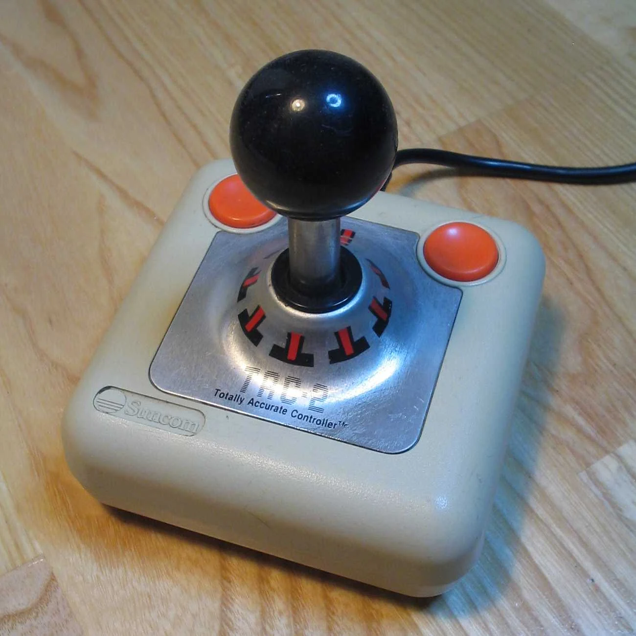 Oryginalny joystick Suncom TAC-2 w kremowej obudowie, czarna kula na chromowanym trzonie, dwa pomarańczowe przyciski i plakietka Totally Accurate Controller